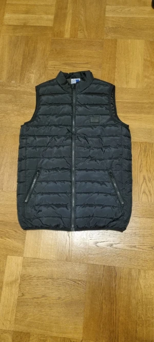 Svart dunväst från Jack & Jones - Svart quiltad dunväst från Jack & Jones med ståkrage och två fickor med dragkedja. Västen har en stilren look med horisontella sömmar och hel dragkedja framtill. Perfekt att slänga över en hoodie för extra värme under höst och vinter.