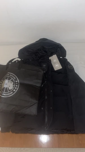 Canada Goose Black Label - Säljer en svart dunjacka från Canada Goose med huva och klassisk logga på ärmen. Jackan har dragkedja framtill, stora fickor och en clean, stilren look. Perfekt för kalla dagar och riktigt snygg till streetwear. Märkestaggar sitter kvar. Vid frågor eller funderingar, skicka DM! Priset är inte hugget i sten!