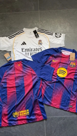 Real Madrid och Barcelona matchtröjor - Säljer tre snygga fotbollströjor: en vit Real Madrid-tröja från Adidas med svarta och gula detaljer, samt två Barcelona-tröjor från Nike med klassiska blå och röda ränder, gul sponsorlogga och klubbmärke. Alla har korta ärmar och är gjorda i lätt, ventilerande material.