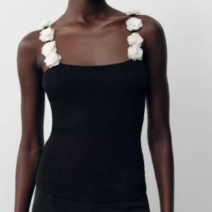 Svart ribbad linnetopp med blommiga axelband - Supersöt svart ribbad linnetopp från Zara med unika axelband täckta av stora, vita tygblommor. Toppen är ärmlös och har en tight passform som framhäver figuren. Perfekt för dig som vill sticka ut med en detaljrik och trendig look.