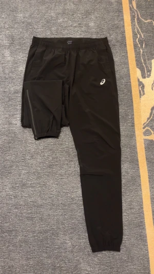Svarta träningsbyxor från Asics M - Svarta träningsbyxor från Asics med elastisk midja och logga på benet. Byxorna har dragkedja vid benslut och är gjorda i lätt polyester som andas. Perfekta för löpning eller gymmet, med smal passform och praktiska detaljer, eller till vardagen. Storlek M. Har Alldrig andvänds bara testas. 