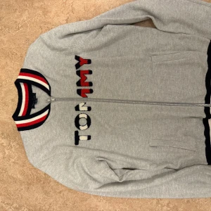 Grå bomberjacka Tommy Hilfiger - Snygg grå bomberjacka från Tommy Hilfiger med broderad logga framtill. Jackan har ribbad krage och muddar i rött, vitt och marinblått, samt dragkedja och två fickor. Perfekt för dig som gillar sportig och clean stil.