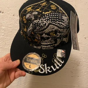 Svart keps med döskalle och glitter - Svart keps från Leader med stora broderade döskallar, guldiga detaljer och glittriga inslag. Kepsen har texten 'Skull' broderad på sidan och framsidan, samt coola mönster med dollar- och dödskallemotiv. Märkets etikett sitter kvar. Bild 1,2 är en keps , bild 3-5 är andra,  135kr/st 