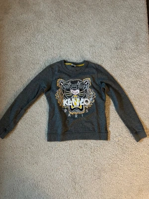 Grå Kenzo sweatshirt med tigertryck - Cool grå sweatshirt från Kenzo i storlek S med det ikoniska broderade tigerhuvudet och Kenzo Paris-text på bröstet. Tröjan har långärmad passform och rund halsring. Tvättlappen är avklippt. Perfekt till vardag!