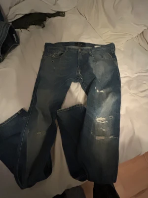 Replay Anbass jeans blå, storlek 34 - Snygga Replay Anbass jeans i blå tvätt med slitna detaljer och distressed look. Klassisk femficksmodell med raka ben och normal passform. Jeansen har tydliga slitningar på framsidan och coola sömmar på bakfickan. Perfekt för dig som gillar en avslappnad streetstil.