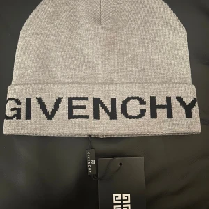 Givenchy Mössa - Snygg grå mössa från Givenchy med bred uppvikt kant och stor svart Givenchy-logga framtill. Tillverkad i mjuk stickad bomull som känns skön mot huvudet. Perfekt för dig som vill ha en clean och lyxig look med tydlig designerstil.
