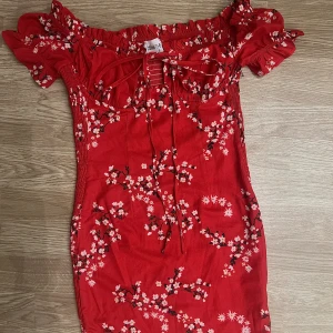 Röd blommig klänning - Supersöt röd kortklänning med blommigt mönster och volangdetaljer vid ärmarna. Klänningen har snörning framtill och smockad rygg för en bekväm passform❤️
