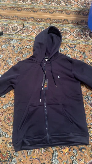 Mörkblå hoodie från Polo Ralph Lauren - Snygg mörkblå hoodie från Polo Ralph Lauren med dragkedja, huva och snörning. Tröjan har en liten broderad logga på bröstet och praktiska fickor framtill. Perfekt för chill dagar och streetstyle.