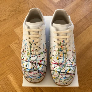 Maison Margiela sneakers med färgstänk - Maison Margiela sneakers i vitt skinn med coola färgstänk i blått, rött, gult, grönt och svart. Modellen har beige snörning, rund tå och en platt gummisula i brunt. Snyggt konstnärligt mönster som sticker ut och ger en unik vibe.