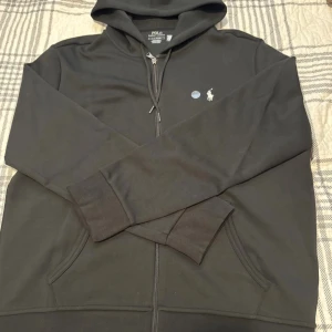 Svart hoodie från Polo Ralph Lauren - Svart hoodie från Polo Ralph Lauren med broderad logga på bröstet. Tröjan har huva med snörning, dragkedja framtill och två fickor. Materialet är mjuk bomull och passformen är normal. Perfekt för chill dagar och streetwear.