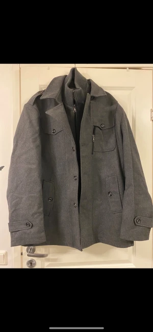 Grå rock med stickad krage - Snygg grå rock med klassisk design och stickad krage. Jackan har både knappar och dragkedja framtill, samt flera fickor med knappstängning. Materialet är ullblandning och insidan har en mjuk stickad del som ger extra värme. Perfekt för kyliga dagar. Kragen är avtagbar.  Jackan är helt nyoch kostar 999kr nypris. 