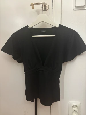 Svart topp från Gina  - Svart topp från Gina Tricot med djup v-ringning och korta, lite fladdriga ärmar. Toppen har knytband i midjan som ger en snygg siluett. Mjuk och skön bomullskvalitet, perfekt att styla med jeans eller kjol. Enkel och stilren design.