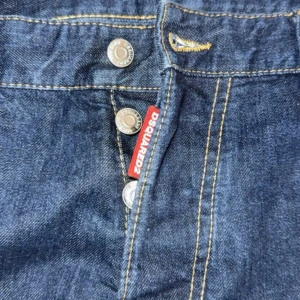 DSQUARED2 ICON jeans blå, strl 52 - Snygga blå jeans från DSQUARED2 med gul ICON-tryck på bakfickan och klassisk DSQUARED2-lapp i svart skinn. Jeansen har synliga knappar framtill, kontrastsömmar och röd DSQUARED2-tag vid gylfen. Perfekt för dig som vill sticka ut med en cool streetstil.