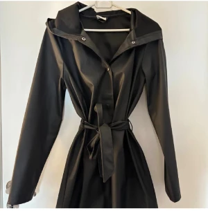 Svart regnjacka med huva och bälte - Svart regnjacka med lång ärm, huva och knytbälte i midjan. Jackan har knappar framtill och är tillverkad i ett slätt, vattenavvisande material. Perfekt för regniga dagar och har en stilren, minimalistisk look.