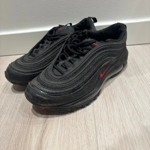 Nike Air Max 97 svarta med röda detaljer - Säljer ett par svarta Nike Air Max 97 med röda detaljer och klassisk vågig design. Skorna har snörning, platt sula och synlig Air-enhet i sulan. Materialet är en mix av syntet och textil. Perfekta för dig som gillar streetwear och sportig stil.