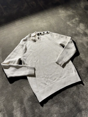 Beige stickad tröja med knappar - Snygg beige stickad tröja från GAP med rund hals och fyra svarta knappar på axeln. Tröjan har ribbade muddar vid hals, ärmslut och nederkant. Perfekt för dig som gillar enkel och stilren design med en liten twist.