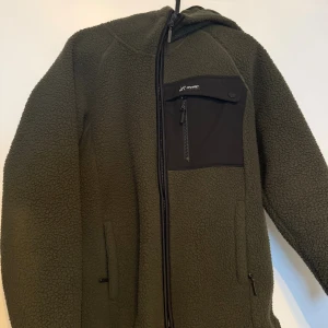 Grön fleecejacka RevolutionRace M - Säljer en grön fleecejacka från RevolutionRace i dammodell, storlek M. Jackan har huva, svart bröstficka med dragkedja och knapp, samt två sidofickor med dragkedja . Snygg, loose passform och långärmad. Använd fåtal gånger 