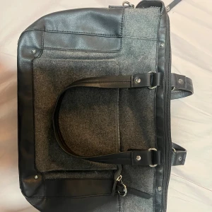 Grå och svart datorväska med dragkedja - Snygg datorväska i grå filt och svarta skinn-detaljer. Väskan har två handtag, en stor ficka med dragkedja på framsidan och metallbeslag. Perfekt för att bära laptop och andra prylar till skolan eller jobbet.