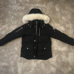 Moose Knuckles jacka - Svart jacka från Moose Knuckles med fluffig vit päls runt huvan. Jackan har långa ärmar med dragkedjor, flera fickor med knappar och snygga silverdetaljer. Perfekt för kalla dagar med sin tjocka och quiltade design. Stilsäker look med coola detaljer.