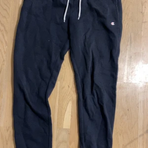 Svarta mjukisbyxor från Champion - Svarta mjukisbyxor från Champion med vit snörning i midjan och klassisk logga på vänster höft. Byxorna har en avslappnad passform och är tillverkade i mjukt bomullsmaterial, perfekta för chill dagar eller träning.