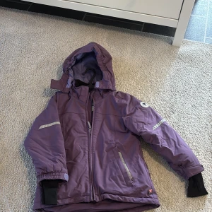 Lila vinterjacka från Polarn O. Pyret - Säljer en lila vinterjacka från Polarn O. Pyret med huva, reflexdetaljer och dragkedja. Jackan har fleecefoder, muddar vid ärmslut och är tillverkad i slitstark, återvunnen polyester. Perfekt för kalla dagar och skyddar mot både vind och regn.
