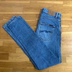 Blå Nudie Jeans straight fit - Säljer ett par klassiska blå jeans från Nudie Jeans med raka ben och normal passform. Jeansen har fem fickor, snygga kontrastsömmar och Nudie-loggan på bakfickorna. Tillverkade i slitstarkt bomullsjeansmaterial och passar perfekt till en avslappnad stil.
