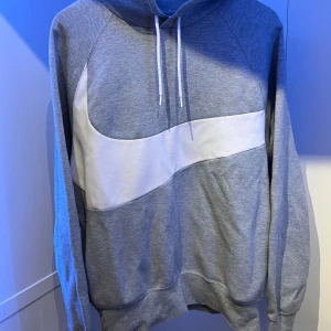 Grå Nike hoodie med vit swoosh - Snygg grå hoodie från Nike med stor vit swoosh som går runt hela tröjan. Tröjan har huva med snörning och långa ärmar. Tillverkad i mjuk bomullsblandning, perfekt för chill eller streetwear. Klassisk och stilren design med broderad text på ryggen.