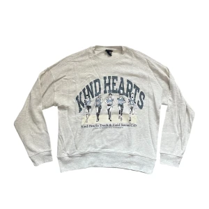 Grå sweatshirt - Ljusgrå sweatshirt med tryck av löpare och texten 'Kind Hearts Track & Field Social Club' framtill. Tröjan har rund hals, långa ärmar och är tillverkad i mjuk bomullsmix. Perfekt för en sportig och avslappnad stil.