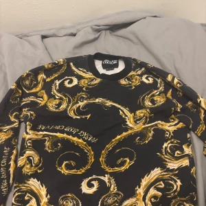 Svart och guld Versace Jeans Couture tröja - Säljer en svart långärmad tröja från Versace Jeans Couture med galet snyggt barockinspirerat mönster i guld och vitt över hela plagget. Tröjan har rund hals och ribbade muddar. Perfekt för dig som vill sticka ut med en lyxig och unik stil. Ny pris 3700kr 800kr om man möts i Enköping kommun 