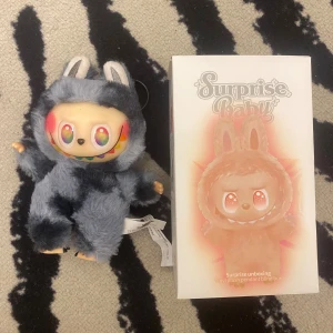 Surprise Baby Vinyl Plush Pendant Blind Box - Unik samlarfigur från Surprise Baby, med färgglad vinylansikte och mjuk plyschdräkt. Produkten är i nyskick och levereras med originalförpackning. Perfekt för samlare eller som present. Inga synliga defekter eller slitage.