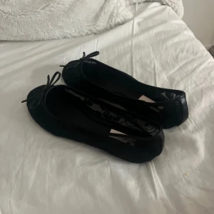 Svarta ballerinaskor med spetsdetaljer - Säljer ett par svarta ballerinaskor från H&M med fina spetsdetaljer och rosett framtill. Skorna har rund tå och platt sula, vilket gör dem både stilrena och bekväma. Perfekta att matcha med jeans eller klänning för en trendig look.
