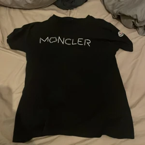 Svart Moncler t-shirt med logga - Svart t-shirt från Moncler med vit logotyp framtill och klassisk Moncler-patch på ärmen. T-shirten har en enkel, stilren design och är kortärmad. Perfekt för dig som gillar exklusiva streetwear-vibbar.