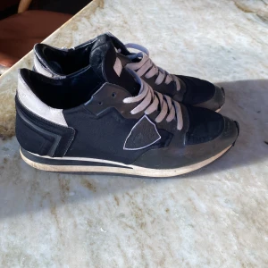 Svarta sneakers från Philippe Model - Snygga svarta sneakers från Philippe Model med vita detaljer och snörning. Skorna har en klassisk siluett med rund tå och platt sula. Ovandelen är i textil och skinn, med märkets logga på sidan. Perfekta för dig som gillar stilrena och sportiga skor.