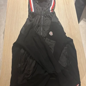 Svart Moncler vindjacka med huva - Svart vindjacka från Moncler med huva och dragkedja framtill. Jackan har detaljer i blankt syntetmaterial upptill och på huvan, samt Moncler-logga på ärmen. Röd och vit rand längs insidan av kragen ger en sportig touch.