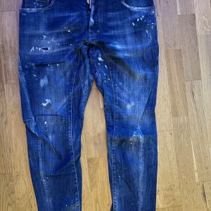 Blå jeans från Dsquared2 med slitningar - Säljer ett par blå jeans från Dsquared2 med coola slitningar och färgstänk för en edgy look. Jeansen har klassisk femficksdesign och är tillverkade i bomull. Perfekta för dig som gillar streetstyle och vill sticka ut lite extra.