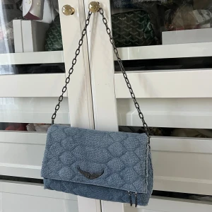 Quiltad blå axelväska från Zadig & Voltaire - Snygg quiltad axelväska i ljusblått tyg från Zadig & Voltaire med metallkedja som axelrem. Väskan har ett coolt vingemblem i silver framtill, dragkedja både på baksidan och insidan samt fransad kant. Perfekt storlek för det viktigaste. Det medföljer en till rem som är blå. 