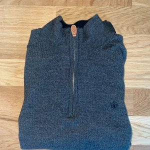 Grå half zip tröja från Morris - Snygg grå stickad tröja från Morris i 100% merinoull. Tröjan har half zip-dragkedja, hög krage och diskret broderad logga på bröstet. Perfekt för lager-på-lager och passar till många olika stilar.