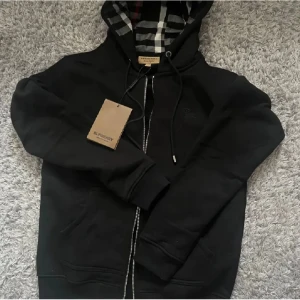 Svart Burberry hoodie med dragkedja - Svart hoodie från Burberry med klassiskt rutigt foder i huvan och broderad logga på bröstet. Tröjan har dragkedja framtill, snörning i huvan och är tillverkad i mjuk bomull. Perfekt för dig som gillar stilrena och exklusiva streetwear-plagg. Helt ny tröja oanvänd 