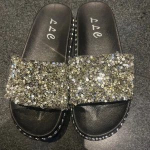 Glittriga svarta sandaler med paljetter - Svarta sandaler med bred rem täckt av glittriga paljetter och stenar. Sulan är platt och dekorerad med små stenar längs kanten för extra bling. Perfekta för dig som vill sticka ut och addera lite glamour till din outfit. Passar 36 också. 