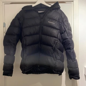 Dunjacka Peak Performance - Säljer en svart dunjacka från Peak Performance med huva och dragkedja. Jackan är tjockt vadderad och har en klassisk pufferstil, perfekt för kalla dagar. Snygg och enkel design med logga på bröstet och praktiska fickor.