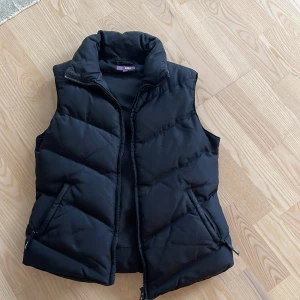 Svart dunväst från Zoul - Svart dunväst från Zoul med hög krage och dragkedja framtill. Västen har två fickor med dragkedja. Perfekt att slänga över en hoodie för en chill look.