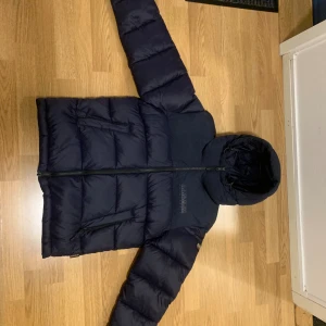 Mörkblå pufferjacka från Napapijri - Säljer en mörkblå pufferjacka från Napapijri med stor logga på ryggen och mindre logga på bröstet. Jackan har huva, dragkedja framtill och två snedställda fickor med dragkedja. Perfekt för kalla dagar och riktigt snygg streetstil. (Använd i knappt 1 månad)
