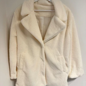 Krämvit teddyjacka med stora slag - New, never used before. Supermjuk och fluffig teddyjacka i krämvitt med stora slag och dolda tryckknappar framtill. Jackan har två fickor och är helfodrad för extra komfort. Perfekt för dig som vill ha en stilren och cozy look under kyliga dagar.