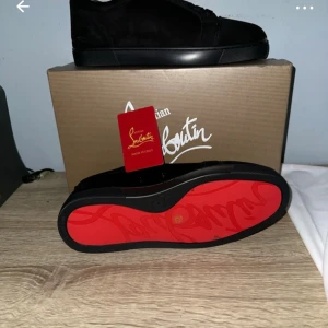 Christian Louboutin svarta sneakers mocka - Snygga svarta sneakers från Christian Louboutin i mocka med klassisk röd sula och broderad logga på hälen. Skorna har rund tå, snörning och platt sula. Perfekta för dig som vill sticka ut med exklusiv design och lyxig känsla.