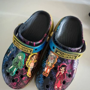 Färgglada Crocs Rainbow High - Unika Crocs med Rainbow High-tema, dekorerade med färgglada figurer och graffiti-liknande mönster. Skorna har svart bas med inslag av blått, gult och rosa samt glittrig guldkant runt sulan. FÖR BARN 