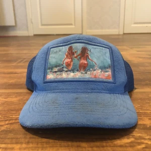 Blå keps med mesh och konstmotiv - Säljer en blå keps med mesh på sidorna och baksidan för extra ventilation. Framsidan har ett konstnärligt tryck av två personer som springer mot havet. Kepsen har böjd skärm och är tillverkad i syntetmaterial.