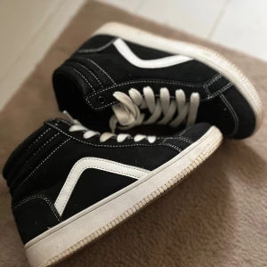 Svarta high-top sneakers med vit detalj - Snygga svarta high-top sneakers från Lejon med vit sula och vit sidodetalj. Skorna har vita snören och kontrasterande vita sömmar som ger en cool look. Ovandelen är i mocka och de har en platt sula och rund tå. Perfekta för dig som gillar streetstyle. Köparen står för frakten 