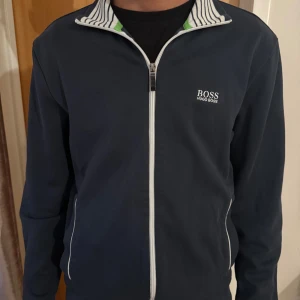 Mörkblå ziptröja från Hugo Boss - Säljer en mörkblå ziptröja från Hugo Boss med vit dragkedja och logga på bröstet. Tröjan har ståkrage med randigt innerfoder i vitt och blått samt gröna detaljer. Långa ärmar och fickor med vita detaljer. Perfekt för en sportig och stilren look.
