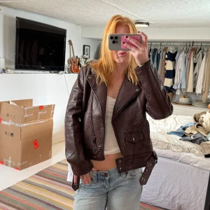 Mörkbrun skinnjacka med bälte - Säljer en mörkbrun skinnjacka i bikerstil med breda slag, dragkedja och bälte i midjan. Jackan har coola detaljer som fickor med lock och knappar samt en croppad passform. Perfekt för dig som vill ha en edgy look.