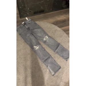 Grå slitna jeans med raka ben - Säljer ett par grå jeans med snygga slitningar vid knäna och raka ben. Jeansen har klassisk femficksdesign och är perfekta för en avslappnad streetstil. Passar dig som gillar en trendig och avslappnad look.